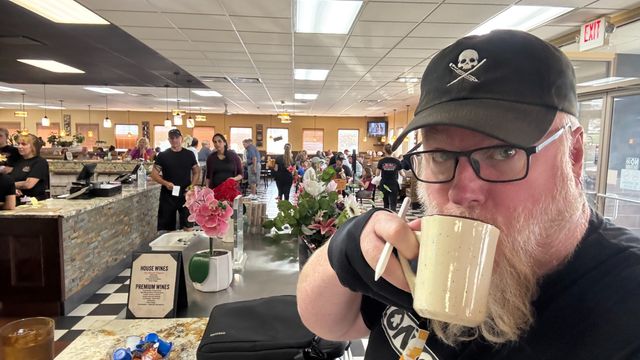 Matt sipping coffee at a local diner. People are milling about behind him and the restaraunt is busy.