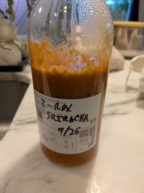 Bottle of homemade sriracha, with a dark orange tone and a scrawled label reading T-Rox Sriracha 9/26
